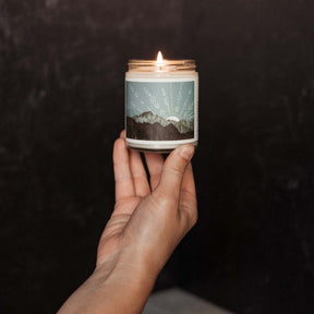 A hand holds up clear glass jar candle with a black lid and mountain landscape artwork. The Peak Jar Candle is hand-poured by Particle Goods in Seattle, WA.