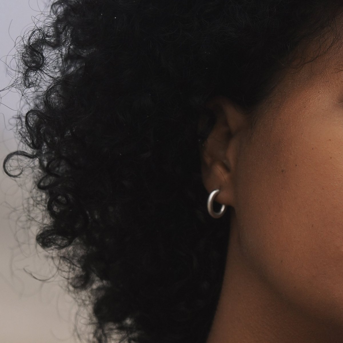 A model wears a smooth silver tone oval shaped hoop with sterling silver earring posts. The Pool Hoops in Silver are designed and handcrafted by Natalie Joy in Portland, Oregon.
