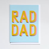 Greeting card reads "RAD DAD" in yellow and pink print against a light blue background. Printed by Banquet Atelier in Vancouver, Canada.