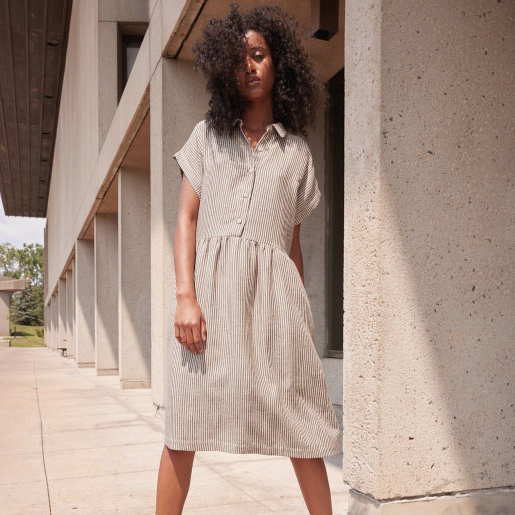 A model wears the flow striped striped dress with short sleeves, collar and buttons up the front. The Reyna dress in Natural & Noir is designed by Eve Gravel and made in Montreal, Canada. 