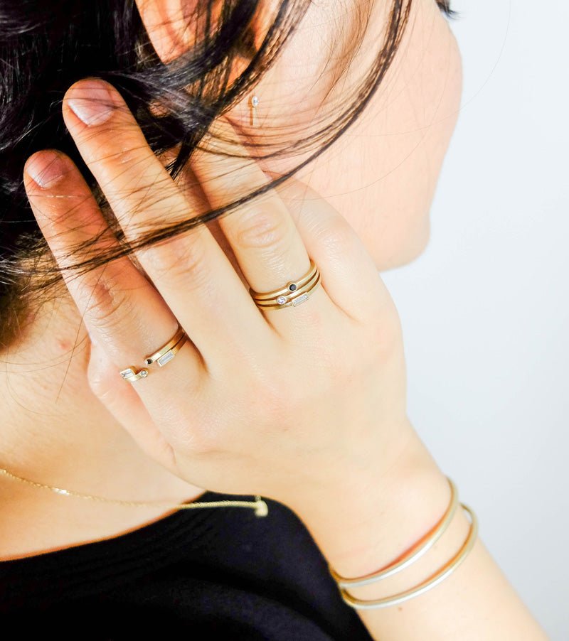 Stacks of betsy & iya's 14k yellow gold and diamond Robur, Nitor, and Manus rings, styled on a model wearing betsy & iya white diamond Navitas earrings and a black shirt.