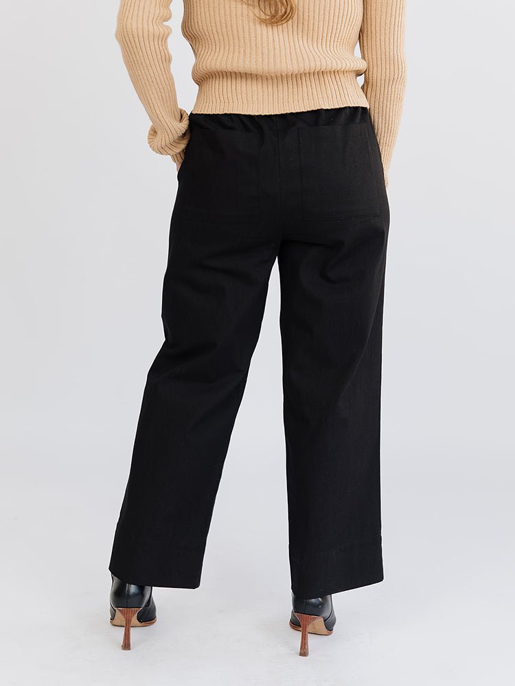 A model shows the back side of a straight leg loose denim pant in black with an elastic waist. The Rosie Pant in Black Denim is designed by Mata Traders and made in India.