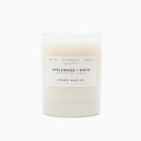 A cylindrical glass candle filled with a soy wax blend and two double cotton wicks. Label reads: "397g/14 oz. Manufactured in Virginia, USA by Sydney Hale Co. Applewood + Birch scented soy candle. Sydney Hale Co. Supporting Animal rescue since 2010."
