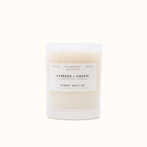 A cylindrical glass candle filled with a soy wax blend and two double cotton wicks. Label reads: "397g/14 oz. Manufactured in Virginia, USA by Sydney Hale Co. Cypress + Cassis scented soy candle. Sydney Hale Co. Supporting Animal rescue since 2010."