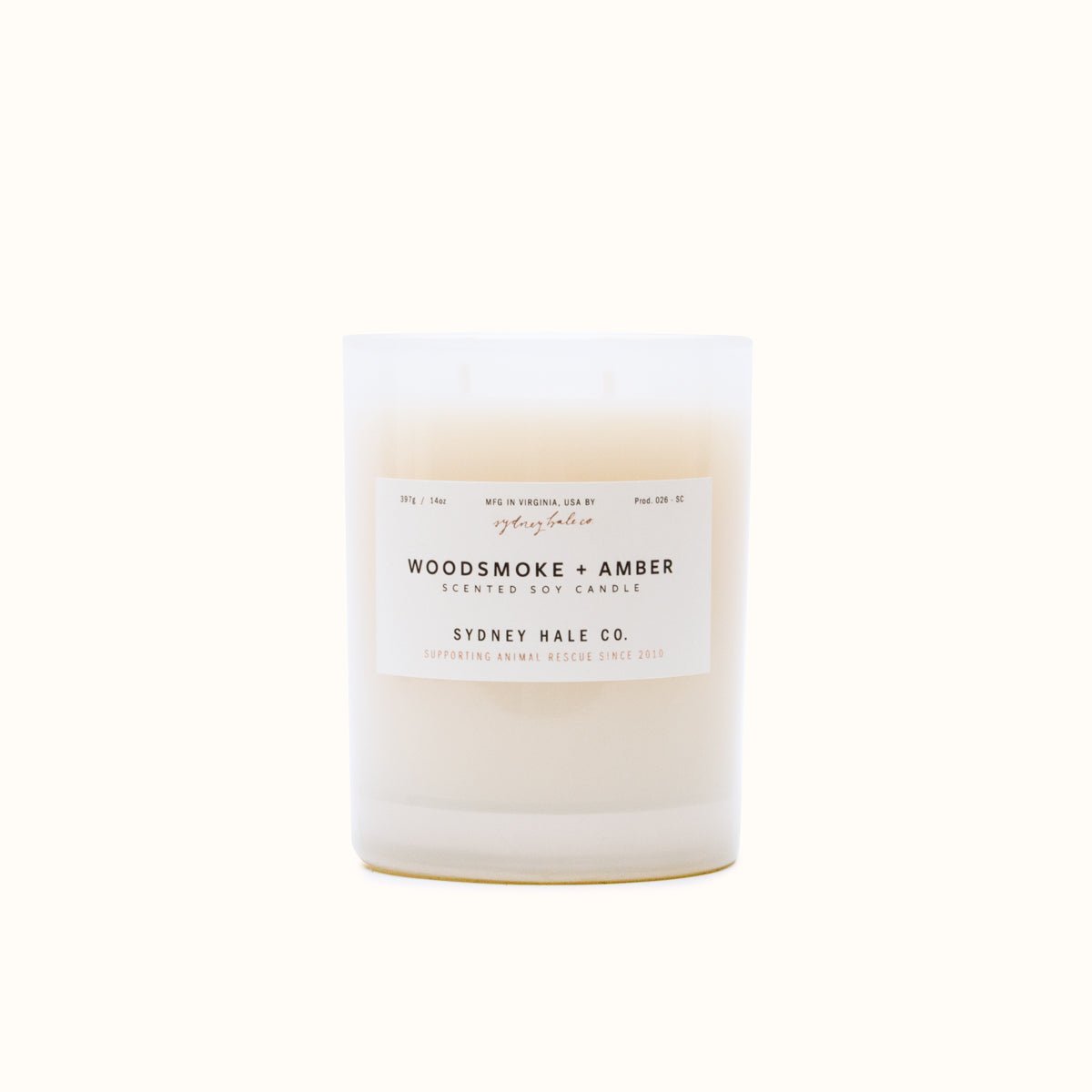 A cylindrical glass candle filled with a soy wax blend and two double cotton wicks. Label reads: "397g/14 oz. Manufactured in Virginia, USA by Sydney Hale Co. Woodsmoke + Amber scented soy candle. Sydney Hale Co. Supporting Animal rescue since 2010."