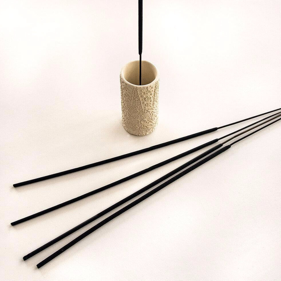 Cream colored cylindrical incense burner with an external embossed pattern. Holds a single stick of black incense. Four sticks of black incense lay in front of holder.  Made by Skeem Design in Philadelphia, PA.