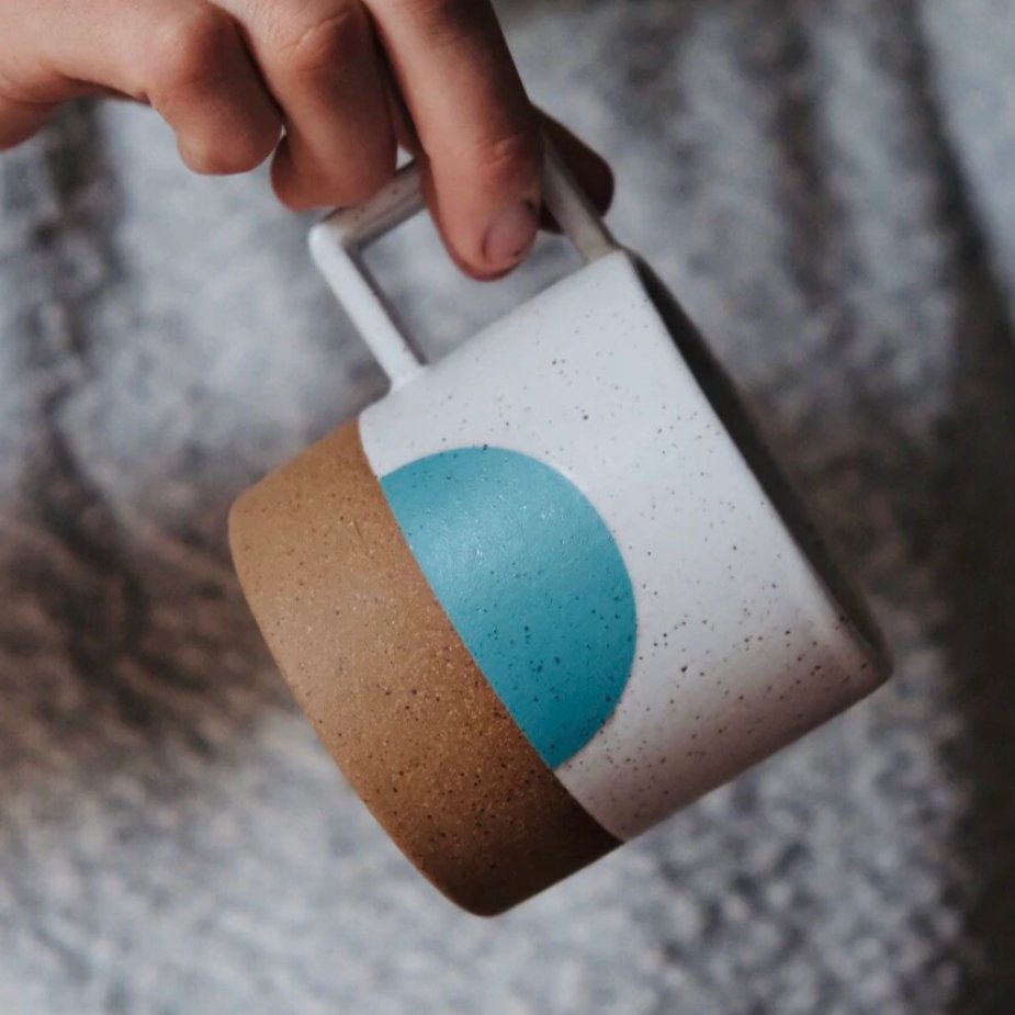 A mug with a white speckled and natural clay finish with a half moon design in a bright teal. The Speckled Moonrise Mug is designed and handmade by Wolf Ceramics in Portland, Oregon.