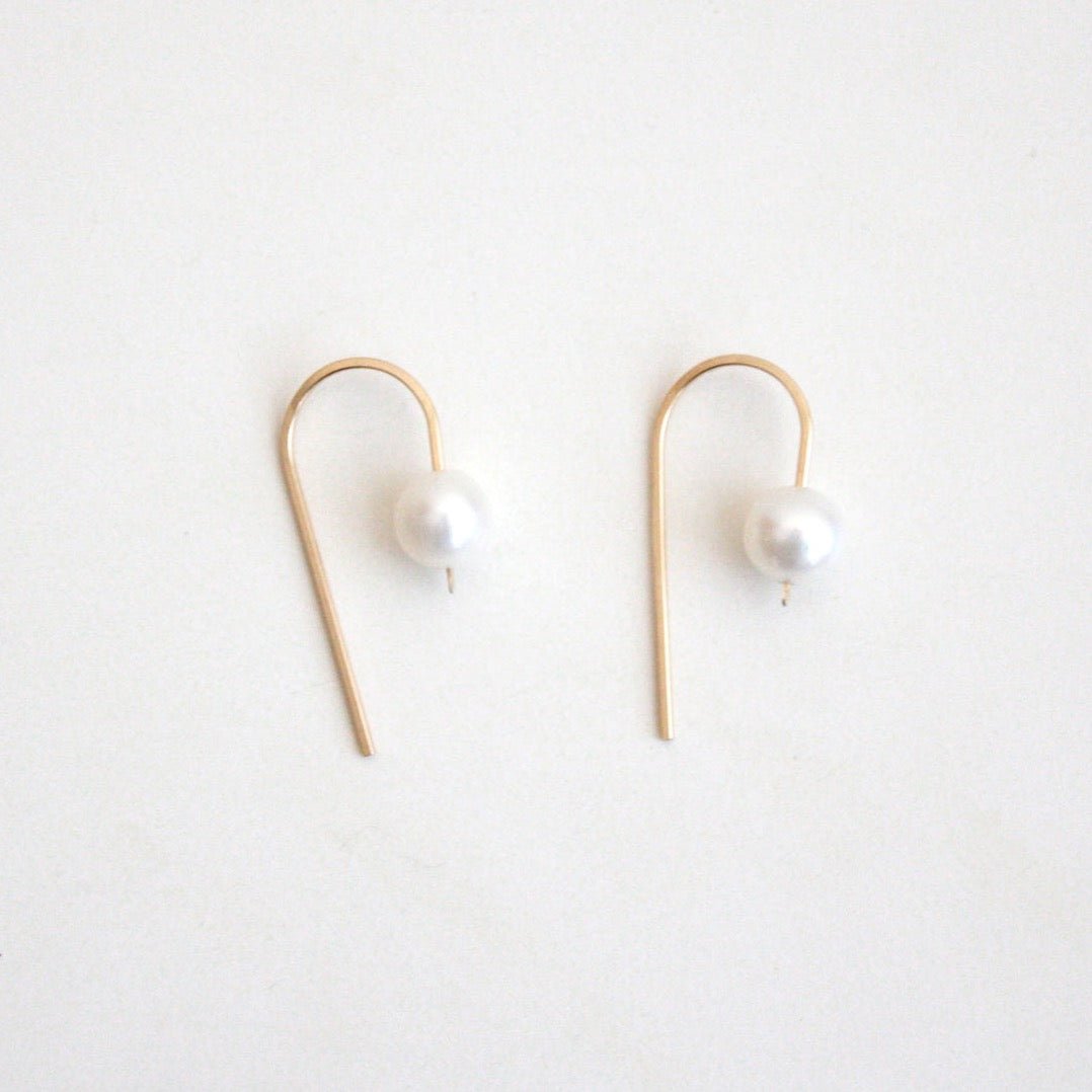 A single gold tone u-shape earring holds a single freshwater pearl. The Short Arc Threader earrings with Large Pearl is designed by Hooks and Luxe and handcrafted in Jackson Heights, NY.
