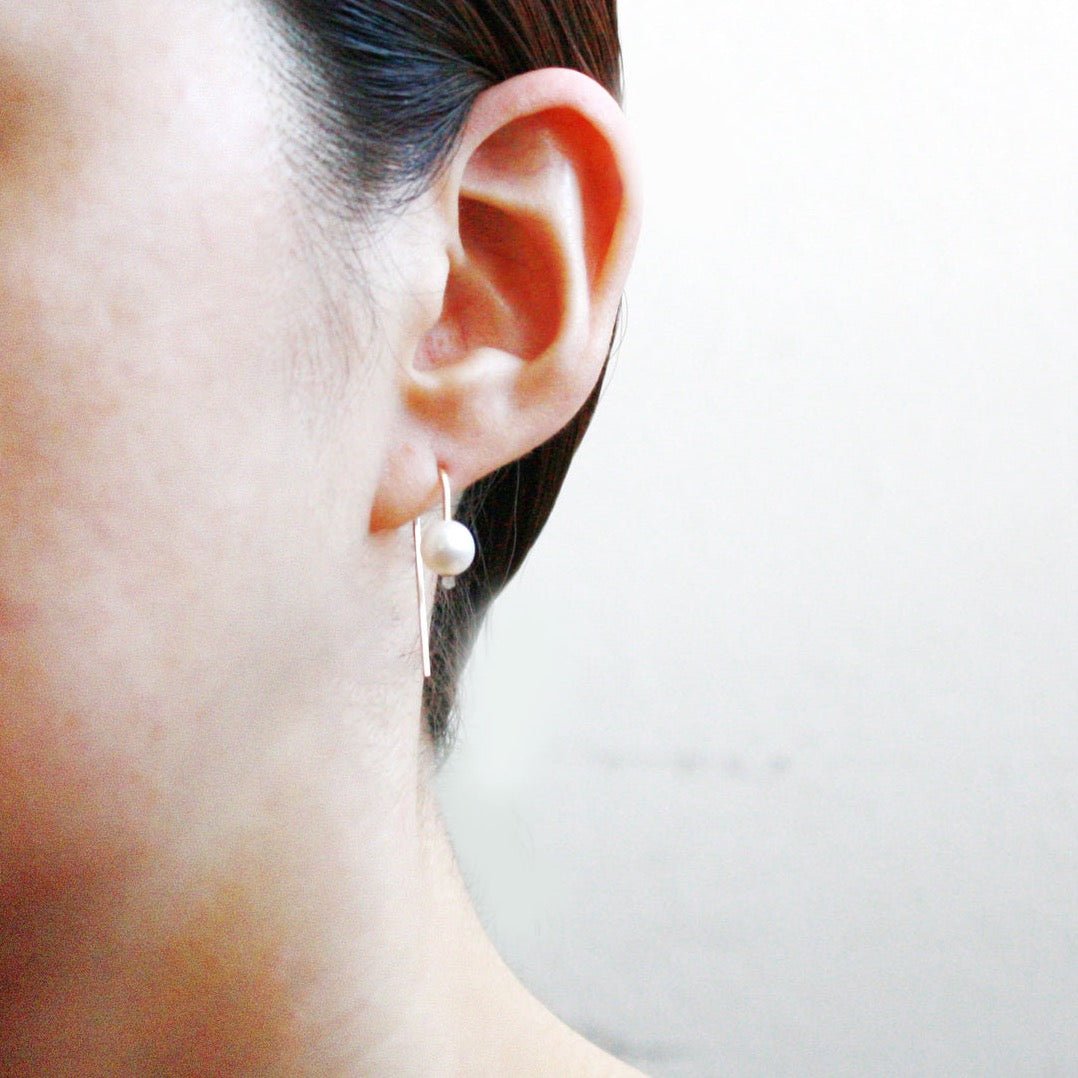 A model wears a gold tone u-shaped earring with a single freshwater pearl. The Short Arc Threader earrings with Large Pearl is designed by Hooks and Luxe and handcrafted in Jackson Heights, NY.