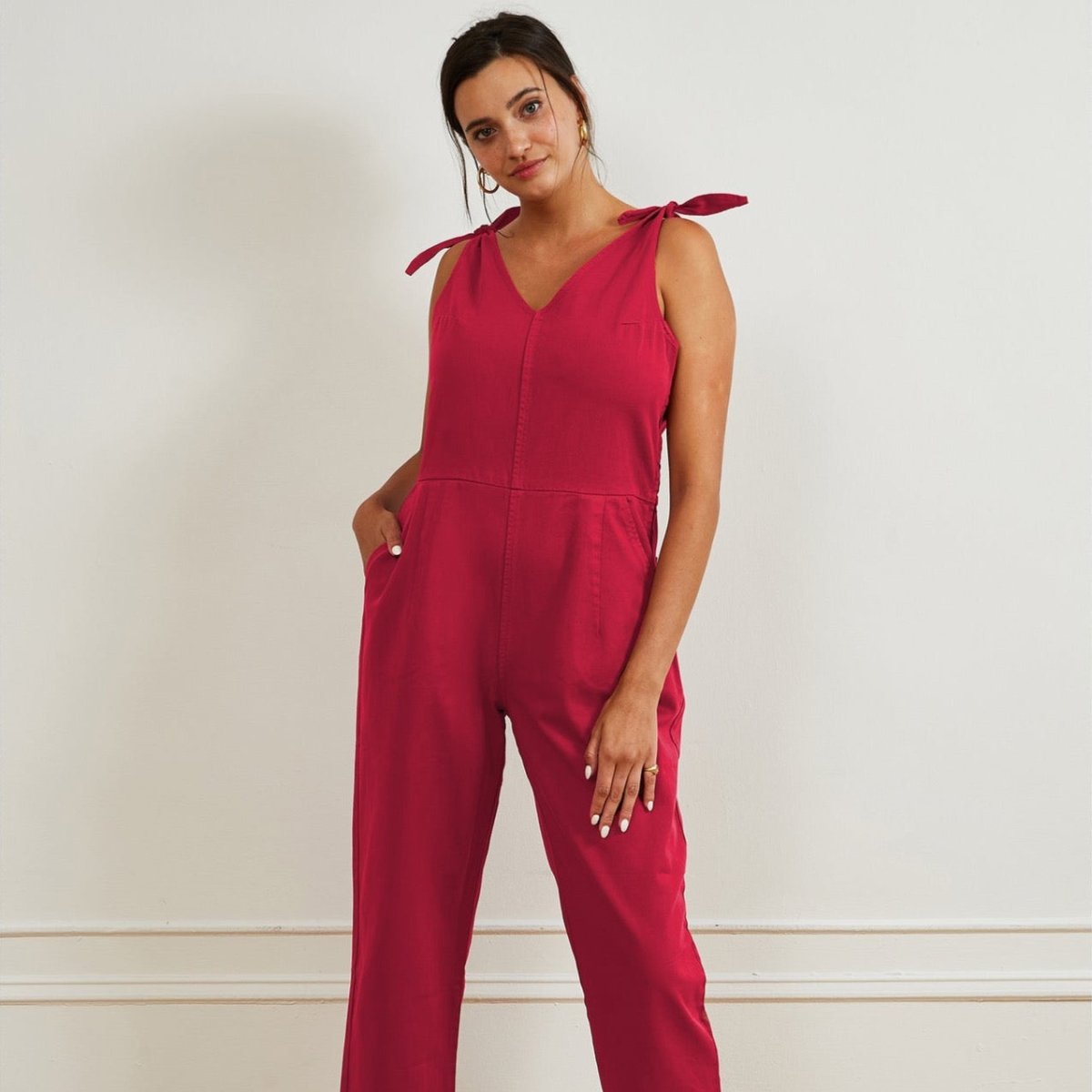 Model wears a bright pink colored jumpsuit with a V-neckline and tie up shoulder straps. The Slate Overalls in Rose are designed by Loup and made in New York City, USA.