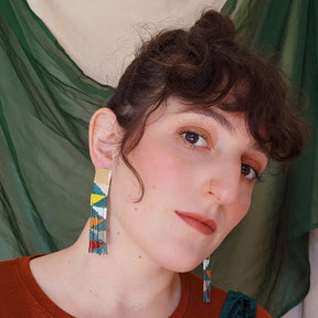 Model wears beaded fringe earrings with a brass square stud. Beads are in emerald, white, yellow, red, grey, orange and salmon. They create a triangular pattern throughout. The Small Kaleidoscope Beaded earrings are designed and handmade by Take Shape Studio in Berkley, California.