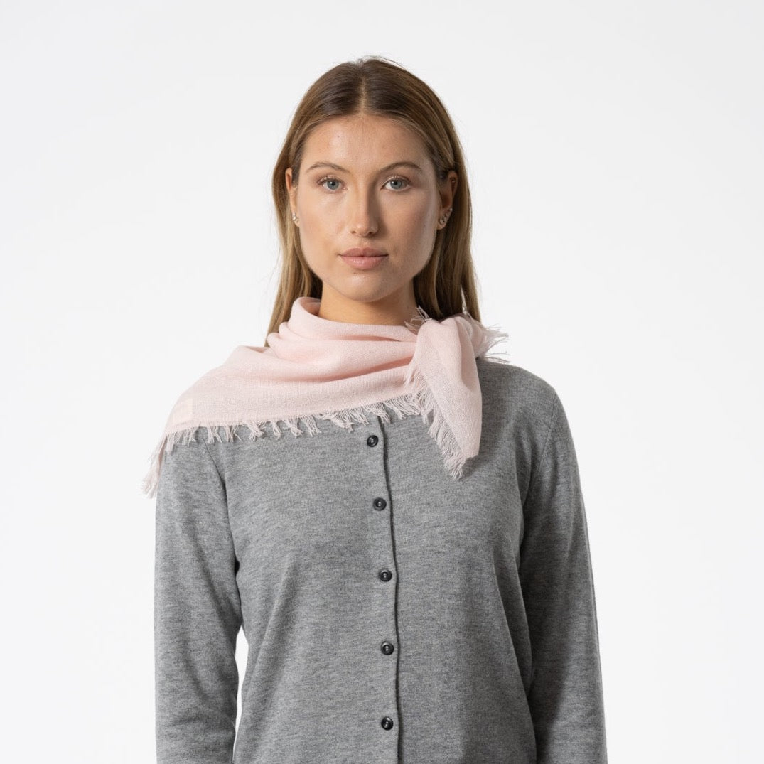 A model wears a square scarf made of 100% Merino wool in a light pink color. The Merino Square Woven Scarf in Blush Pink is designed by Dinadi and handcrafted in Nepal.