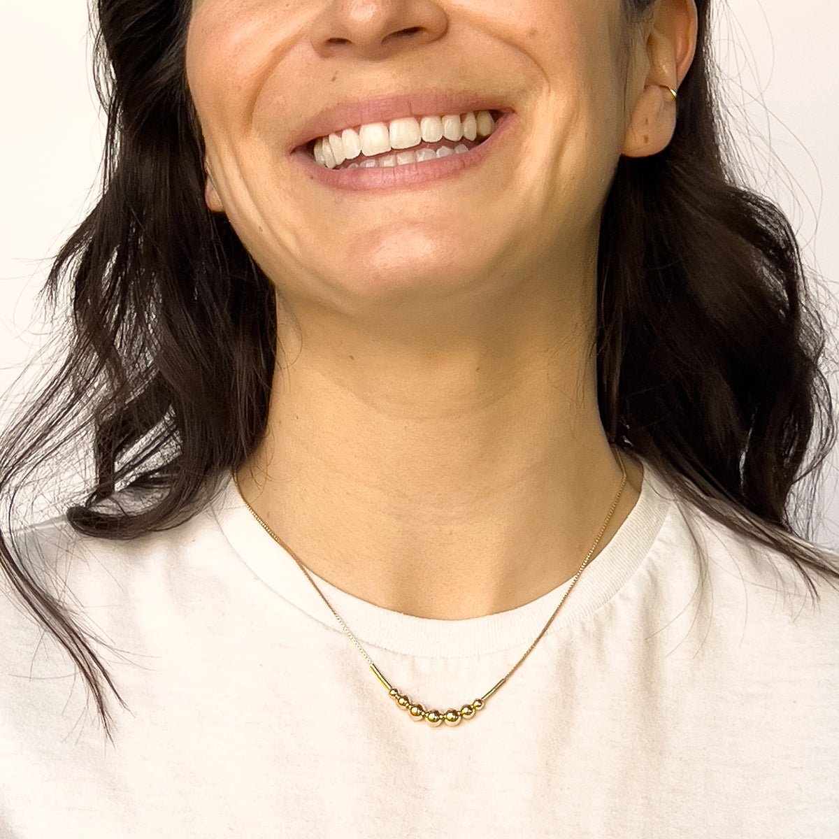 A model wears a gold fill necklace featuring a delicate chain and seven round beads flanked by narrow tube beads. The Strella Necklace is designed and handcrafted in Portland, Oregon.