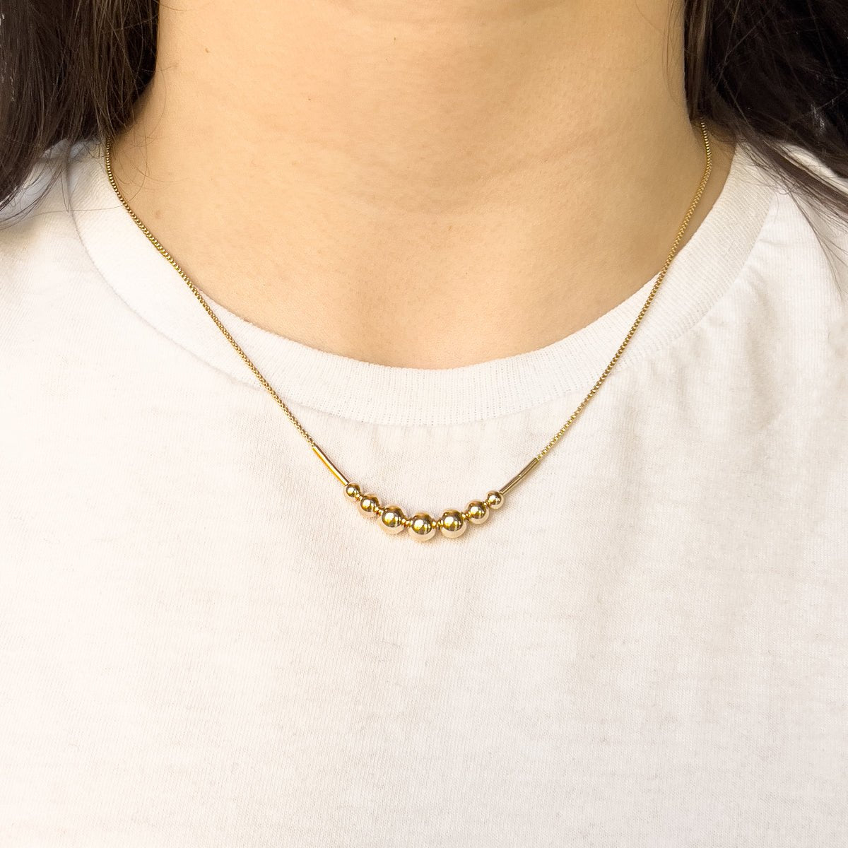 A model wears a gold fill necklace featuring a delicate chain and seven round beads flanked by narrow tube beads. The Strella Necklace is designed and handcrafted in Portland, Oregon.