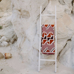 A grey, white, gold, chocolate and black patterned throw displayed on a blanket ladder. The Sunset Mesa Throw is designed by Native American knitwear designer Jennifer Berg and made in New Mexico, USA.