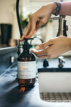Person pumping soap out of an amber bottle by the sink. The soap is from Sydney Hale CO and hand blended and bottled in Richmond, Virginia USA.