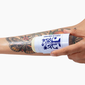 A tattoo balm in a white applicator with a navy blue design held up to a model's tattooed arm. The tattoo balm stick is designed and made by People of Substance in New York, NY.