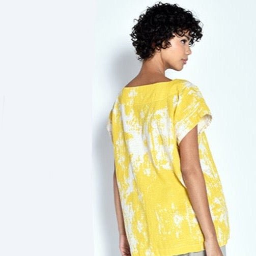 A model shows the backside of a short sleeve tunic with a bright yellow painted design. The Tun in Yellow Bark is designed by UZI NYC and sewn in Brooklyn, New York.