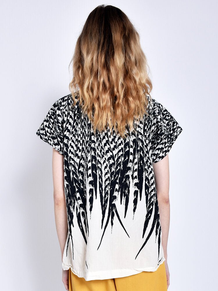 A model shows the back side a short sleeve white top with a black feather design. The Tunic in Cream feather is designed and sewn by Uzi NYC in Brooklyn, New York.