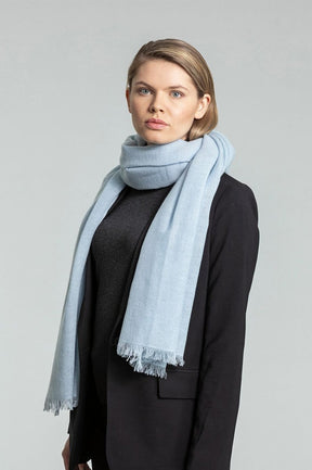 Model shows light blue scarf wrapped around neck. Scarf has frayed edges. The Merino Woven Scarf in Mint Blue is designed by Dinadi and handmade in Kathmandu, India.