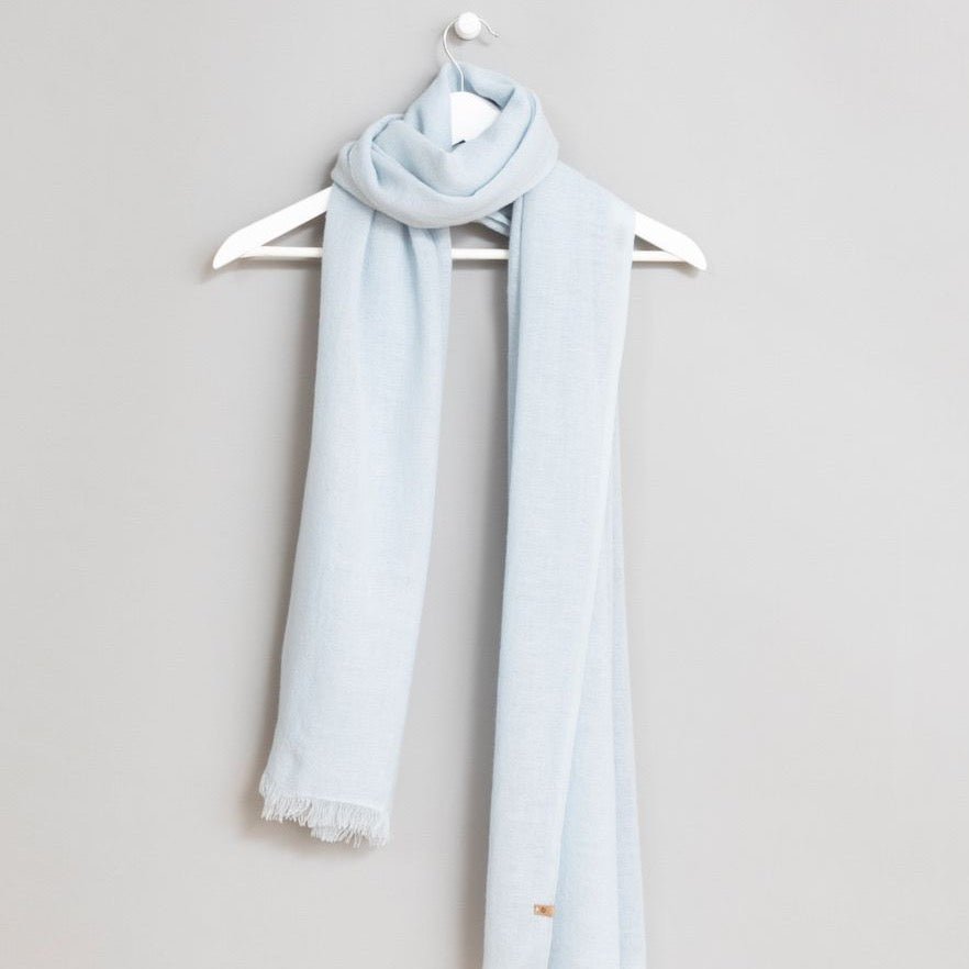 Light blue scarf wrapped around a white hanger. Scarf has frayed edges. The Merino Woven Scarf in Mint Blue is designed by Dinadi and handmade in Kathmandu, India.
