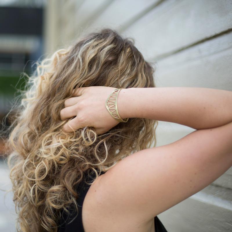 The brass Hernando de Soto Bridge cuff bracelet by betsy & iya, pictured on the wrist of a model with curly, blonde hair. 
