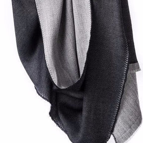 Merino wool scarf hangs in the air. One side is light grey and the other side is a dark charcoal grey or black. Black stitching can be seen on the edges. The Eden Reversible Scarf in Black is from Bloom & Give.