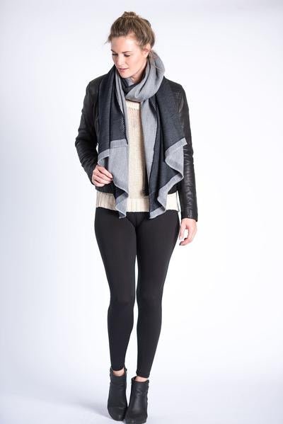 Woman wears a leather jacket and black pants and boots. She has a grey and black scarf wrapped around her neck. The Eden Reversible Scarf in Black is from Bloom & Give.