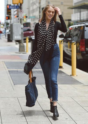 Woman on a city sidewalk wears a black long-sleeved top and blue jeans. She has a black scarf with white dots loose around her neck and a canvas tote bag in her right hand. The Maya Dot Scarf in Black is from Bloom & Give.