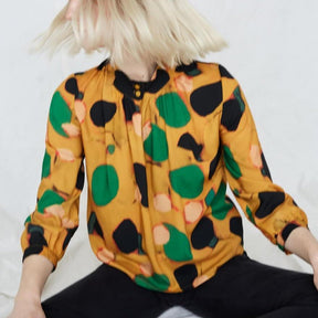 Blonde woman wears yellow blouse with green, pink and black dots. The woman is shaking her hair and sitting crossed-leg in black pants. The Acacia Top in Klimt is from Canadian designer Eve Gravel. Illustrative pattern is a collaboration with artist Catherine D'Amours.