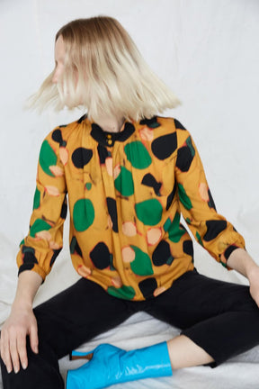 Blonde woman wears yellow blouse with green, pink and black dots. The woman is shaking her hair and sitting crossed-leg in black pants. The Acacia Top in Klimt is from Canadian designer Eve Gravel. Illustrative pattern is a collaboration with artist Catherine D'Amours.