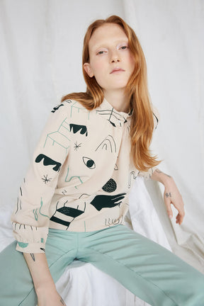 Red-haired woman wearing a cream colored blouse with a black and teal illustrative pattern. The Acacia Top has a buttoned collar and is from Canadian designer Eve Gravel. Illustrative pattern is a collaboration with artist Catherine D'Amours.
