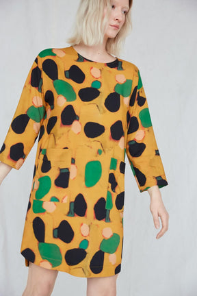 Blonde woman wears a boxy, 3/4 sleeve yellow dress with green, black and pink dots. The Big Trail Dress in Klimt features two large pockets seen in the front, and the dress falls above the woman's knees. This dress is from Canadian designer Eve Gravel. Illustrative pattern is a collaboration with artist Catherine D'Amours.