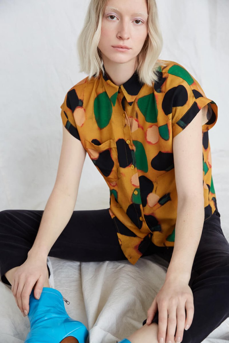 Blonde woman wears a boxy yellow button-down blouse with green, black and pink dots and black pants and bright blue boots. The Caravan Shirt in Klimt is from Canadian designer Eve Gravel. Illustrative pattern is a collaboration with artist Catherine D'Amours.