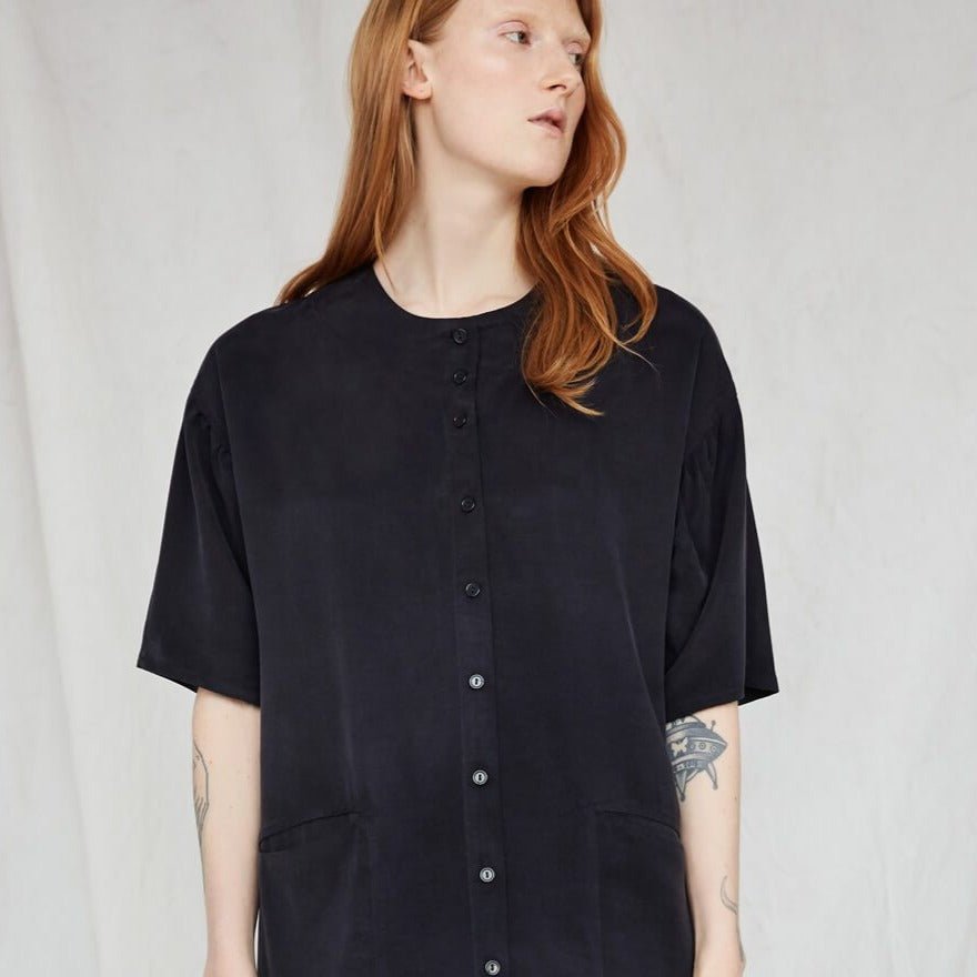 Red-haired model looks to the side while wearing a boxy black button-down dress. The Lullaby Shirt Dress in Black has two side pockets in the front and a round neckline. The dress is from Canadian designer Eve Gravel.