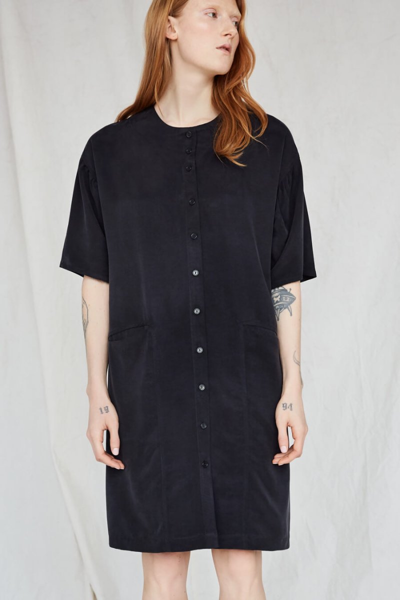 Red-haired model looks to the side while wearing a boxy black button-down dress. The Lullaby Shirt Dress in Black has two side pockets in the front and a round neckline. The dress falls at the model's knee. The dress is from Canadian designer Eve Gravel.