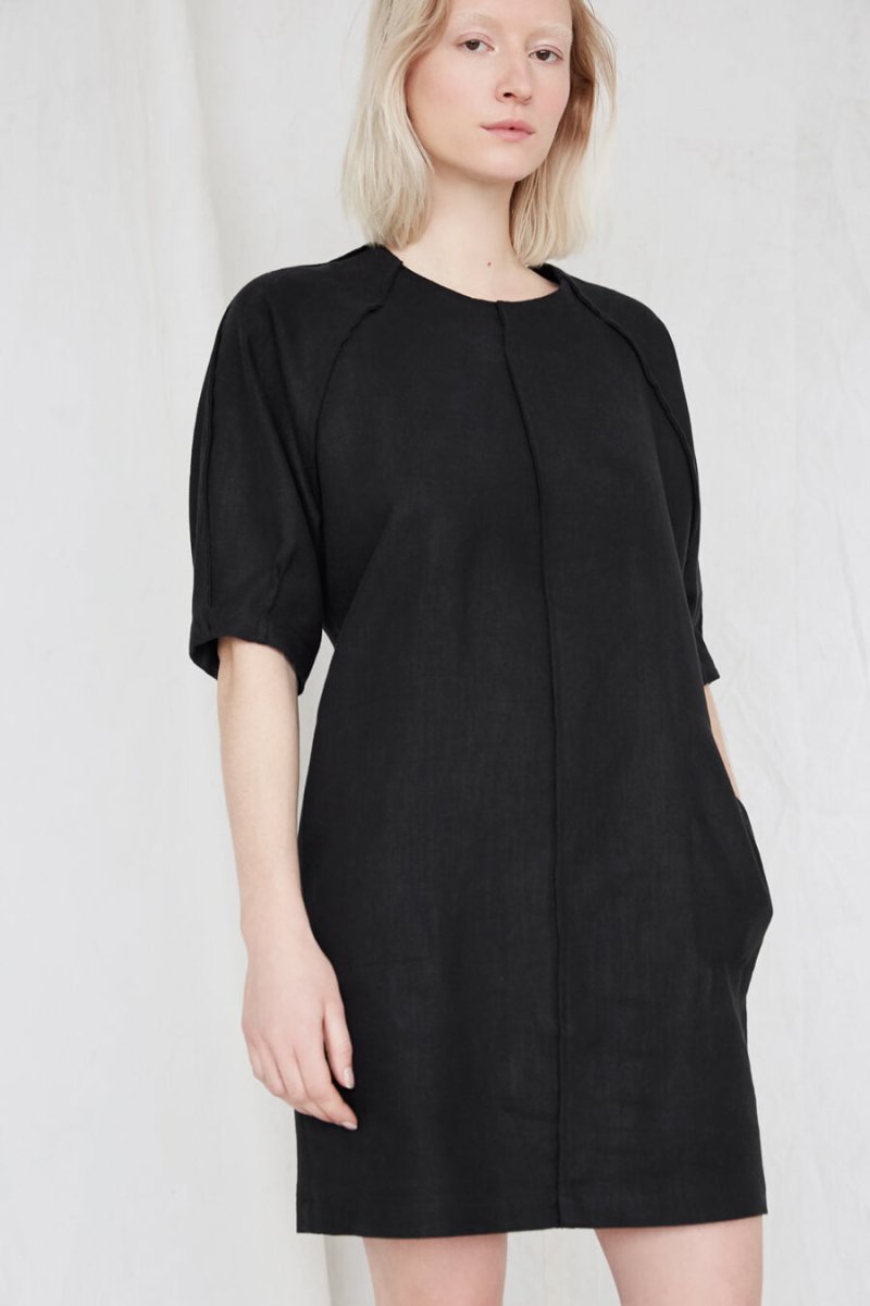 Blonde woman wears a slightly loose black dress with raglan sleeves and has one hand inside a pocket. The Moonless Night Dress in Black features a sleek stitching detail down the front and falls at the model's thighs. The dress is from Canadian designer Eve Gravel.