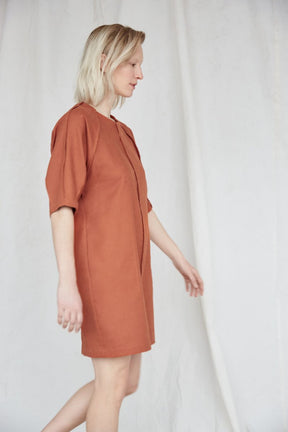 Blonde woman is swinging her arms and wears a slightly loose burnt orange dress with raglan sleeves. The Moonless Night Dress in Burnt Orange features a sleek stitching detail down the front. The dress is from Canadian designer Eve Gravel.
