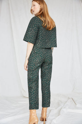 Red-haired woman wears an emerald top and matching pants with intricate jacquard detailing. She is facing the back with her head tilting towards the left. The Night Bird top is from Canadian designer Eve Gravel.
