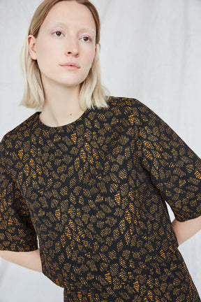 Blonde woman is looking towards the right while wearing an orange and black top with intricate jacquard detailing. The Night Bird top is from Canadian designer Eve Gravel.