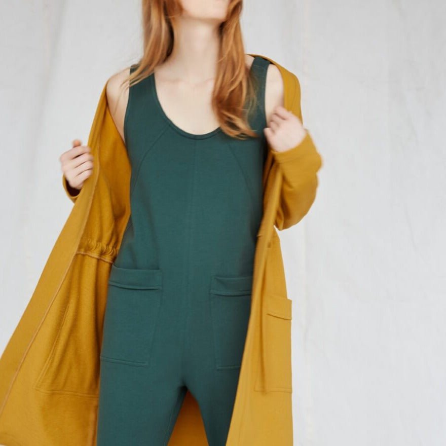 Red-haired woman wears an emerald jumpsuit and a yellow blazer. The Owen Jumpsuit in Spruce is from Canadian designer Eve Gravel.
