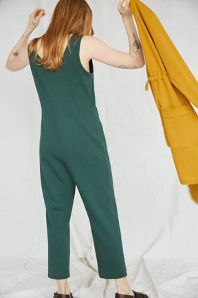 Red-haired woman is facing away and  wears an emerald jumpsuit. In her hand is a yellow coat. The Owen Jumpsuit in Spruce is from Canadian designer Eve Gravel.