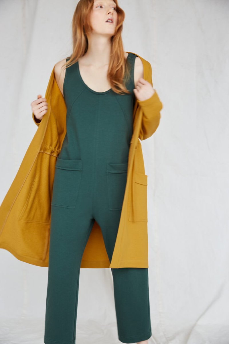 Red-haired woman wears an emerald jumpsuit and a yellow blazer. The Owen Jumpsuit in Spruce is from Canadian designer Eve Gravel.