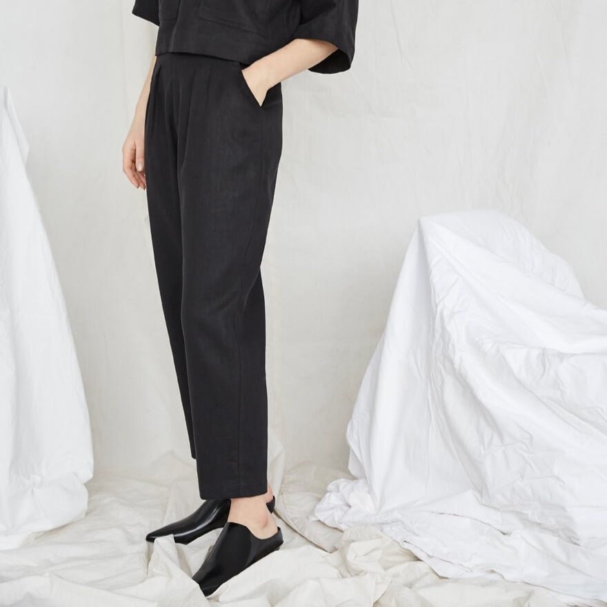 Model is standing with one hand in a pocket and wears a black top and matching pants. The Terre Sauvage Pants in black have pleats in the front and belt loops. The pants fall above the model's ankle and she is wearing black loafers. The pants are from Canadian designer Eve Gravel.