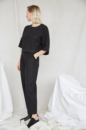 A blonde model is standing while looking away with one hand in a pocket and wears a black top and matching pants. The Terre Sauvage Pants in black have pleats in the front and belt loops. The pants fall above the model's ankle and she is wearing black loafers. The pants are from Canadian designer Eve Gravel.