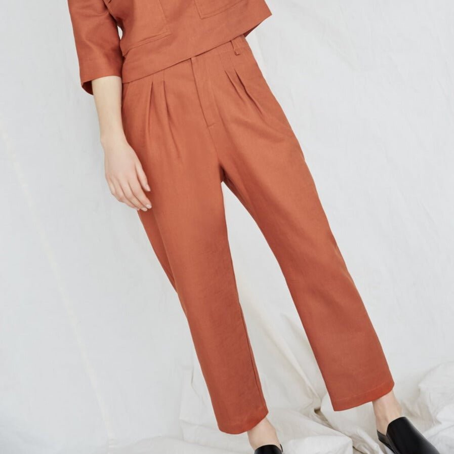 Model is standing at an angle and wears an orange top and matching pants. The Terre Sauvage Pants in burnt orange have pleats in the front and belt loops. The pants fall above the model's ankle and she is wearing black loafers. The pants are from Canadian designer Eve Gravel.