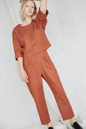 A blonde model is standing at an angle and wears an orange top and matching pants. The Terre Sauvage Pants in burnt orange have pleats in the front and belt loops. The pants fall above the model's ankle and she is wearing black loafers. The pants are from Canadian designer Eve Gravel.