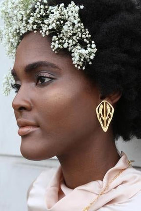 Woman in a profile view wears brass earrings and white flowers in her hair. The Alien Key Post earrings are from designer Lingua Nigra.
