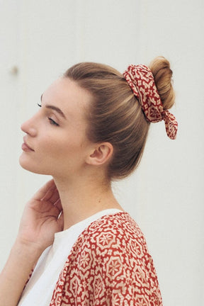 A woman wears a matching red patterned bandana and coverup, while placing her hand on her neck. The Fez Bandana from Maelu is designed in Portland, Oregon and printed by hand in India.