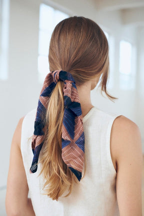 A woman faces away and has her hair pulled back in a ponytail with a bandana. The pink and blue Lola Bandana by Maelu is block printed by hand.
